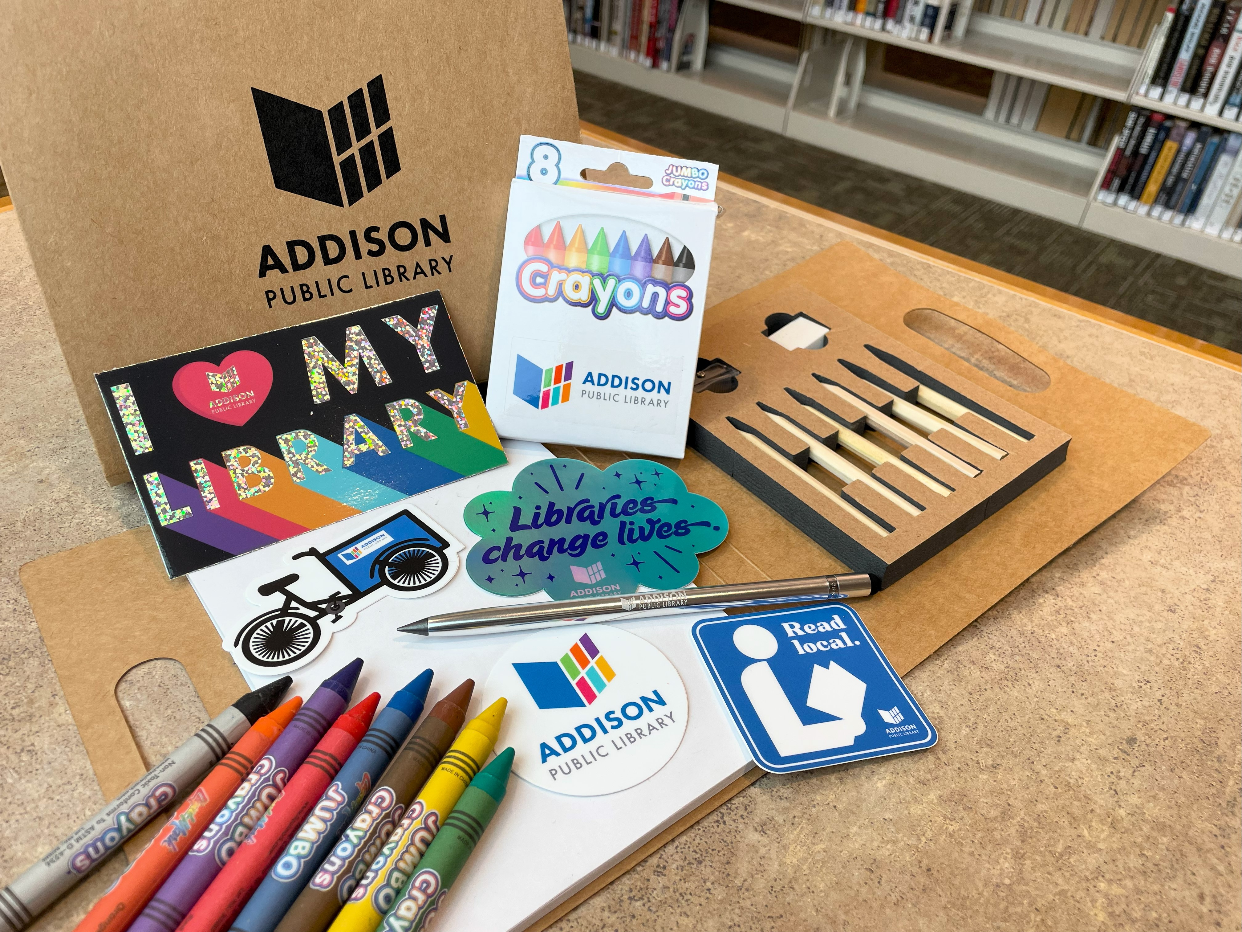 A display of National Library Week giveaways at Addison Public Library. Items include a colored pencil portfolio set, jumbo crayons, a stylus pen, and a variety of colorful library-themed stickers with messages like “I ♥ My Library,” “Libraries Change Lives,” and “Read Local.