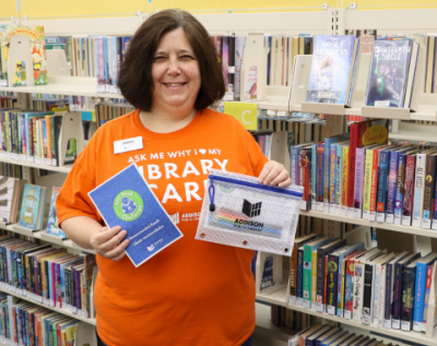 Woman in an orange shirt holding a library branded pouch and a 500 books challenge reading log