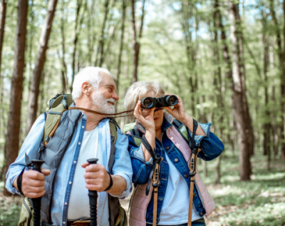 an older couple hiking in the woods with hiking poles and binoculars
