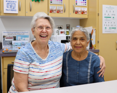 two older women sitting at a table, smiling, one with her arm around the other