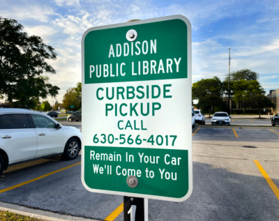 Image of Curbside Pickup parking lot sign. Sign reads, "Addison Public Library. Curbside Pickup. Call 630-566-4017. Remain in your car. We'll come to you."