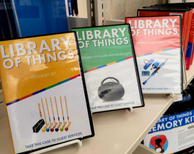 Photo of book shelf with Library of Things display cases. Cases are for the croquet kit, boombox, and fishing pole.
