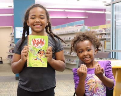 Two children smiling in a library, with one holding a "Yasmin" book.