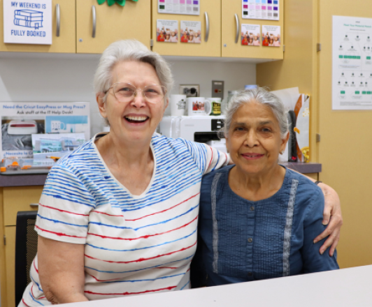 two older women sitting at a table, smiling, one with her arm around the other