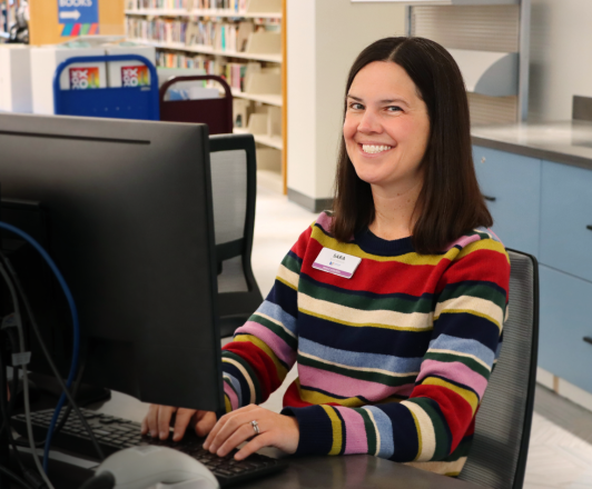 woman with brown hair and striped sweater sitting at a desktop computer smiling
