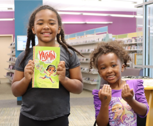 Two children smiling in a library, with one holding a "Yasmin" book.