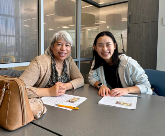 two adults sit at a table smiling with paper handouts and pencils on the table