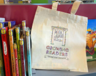 Photo of a tote bag that says "Growing Readers" sitting on a book shelf of picture books. 
