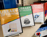 Photo of book shelf with Library of Things display cases. Cases are for the croquet kit, boombox, and fishing pole.