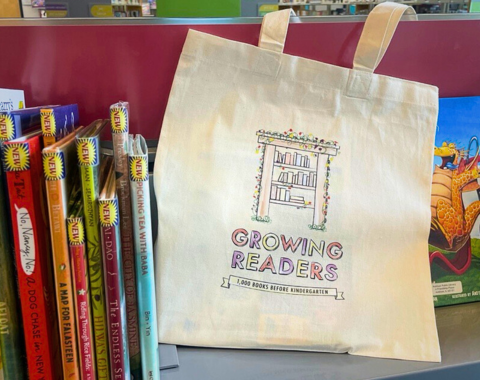 Photo of a tote bag that says "Growing Readers" sitting on a book shelf of picture books. 