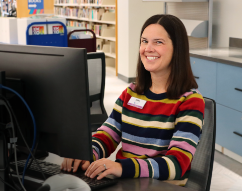 woman with brown hair and striped sweater sitting at a desktop computer smiling