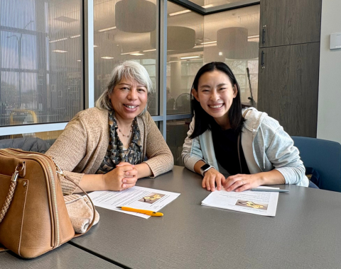 two adults sit at a table smiling with paper handouts and pencils on the table